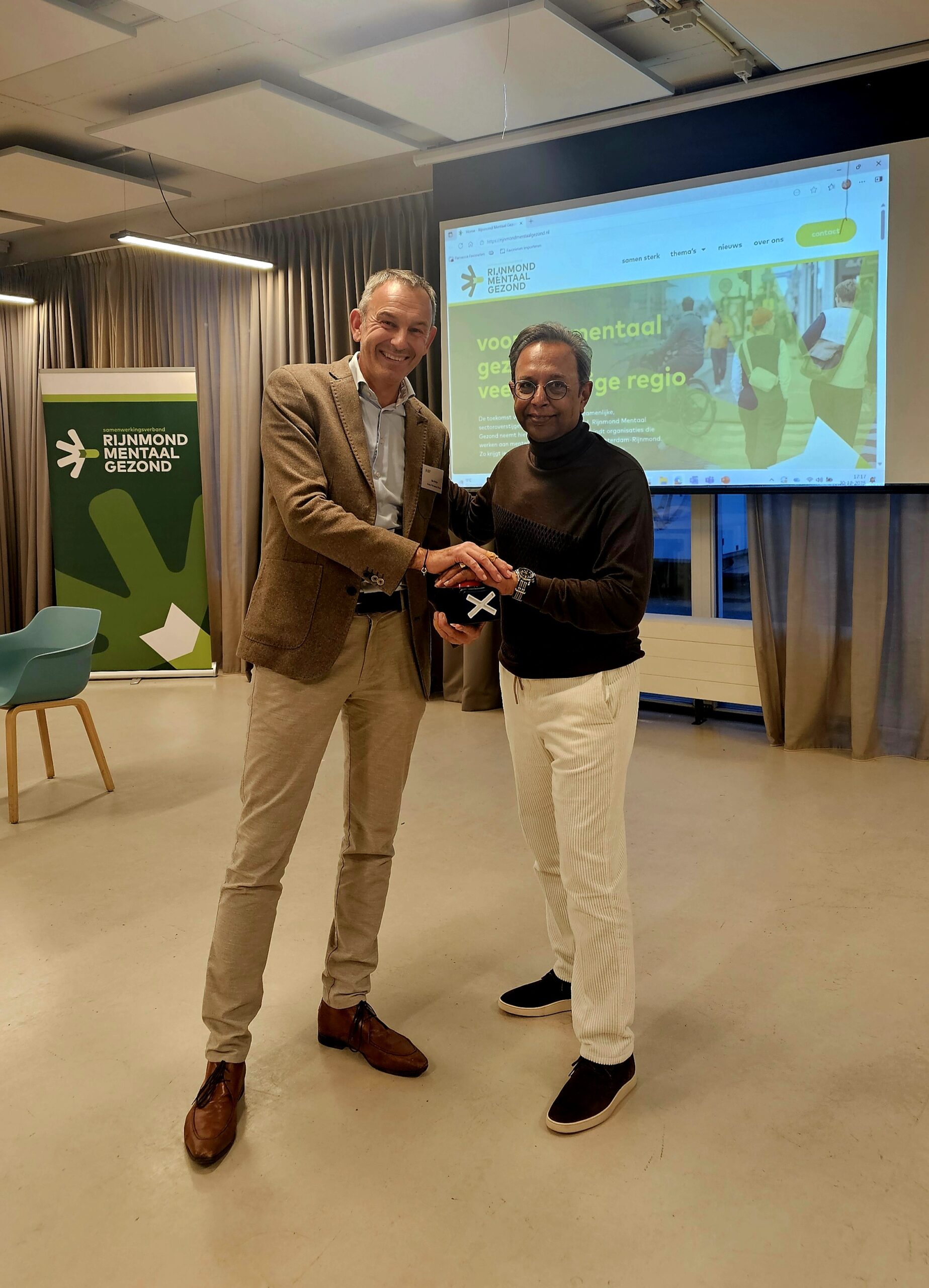 Op de afbeelding staan Bas Molijn, bestuurder Antes en Him Vishnudatt, bestuurder CaleidoZorg. Zij drukken samen op de rode lanceerknop. Op de achtergrond is de website van Rijnmond Mentaal Gezond op een scherm te zien.